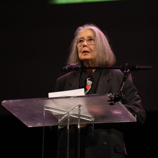 Anne Carson står på en scene ved en klar talerstol med en mikrofon foran en mørk baggrund.