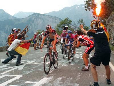 Cykelrytteren Fränk Schleck cykler på Alpe d'Huez under Tour de France 2006. Til højre ses jublende tilskuere. Én af dem med et tændt romerlys i hånden. Bag Schleck ses yderligere cykelryttere - Damiano Cunego og Eddy Mazzoleni.