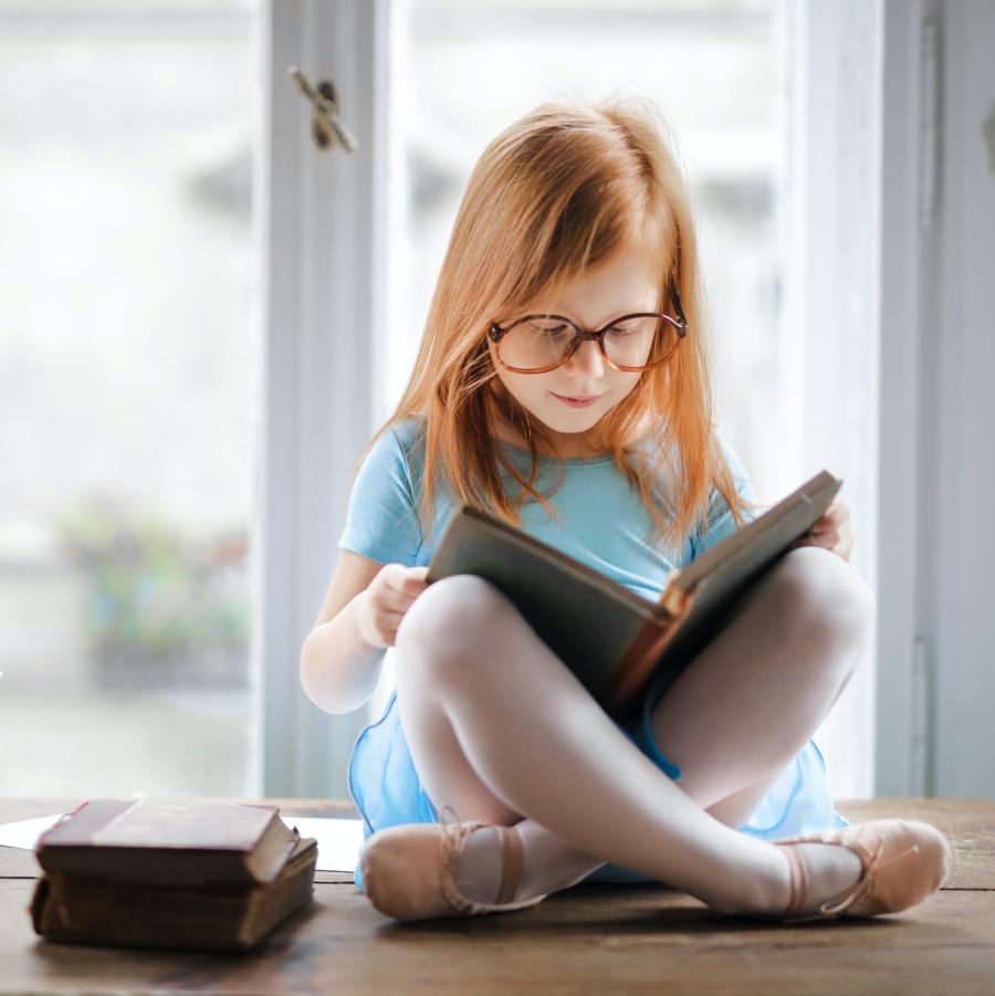 En rødhåret pige med briller, sidder på et bord og læser.